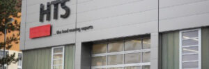 A modern gray building with "HTS" and the slogan "the load moving experts" displayed on its exterior wall.