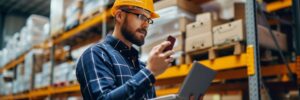 Man working in warehouse. he is using a laptop and holding a mobile phone.